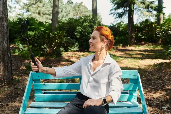 A woman with red hair takes a selfie on a blue bench in a wooded park. — Stock Photo