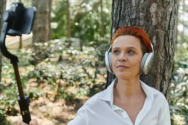 Red haired teacher with nose ring takes forest selfie with headphones and selfie stick — Stock Photo