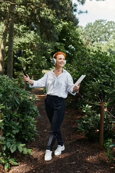 A teacher with red hair and headphones takes a selfie on a campus path. — Stock Photo