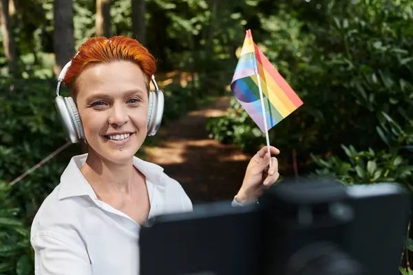 A woman in headphones smiles while holding a rainbow flag in the woods. — Stock Photo