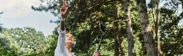 Eine Frau steht in einem Baumhain, hält einen Selfie-Stick in der Hand und blickt in den Himmel. — Stockfoto