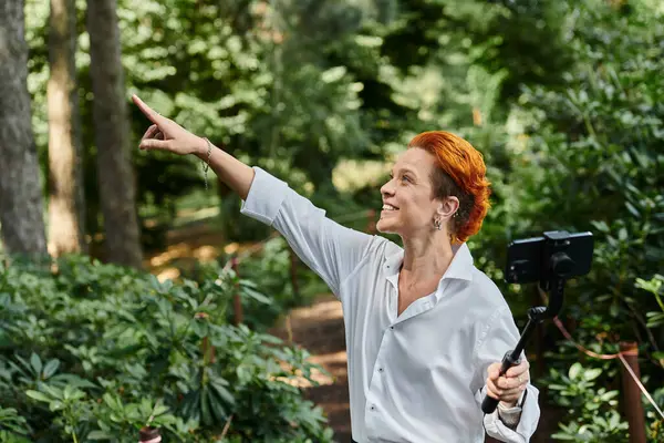 A woman with red hair smiles and films with a selfie stick in a wooded area on campus. — Stock Photo