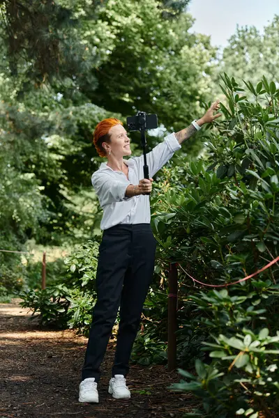 A woman films with her phone on a selfie stick while walking through a lush green campus. — Stock Photo