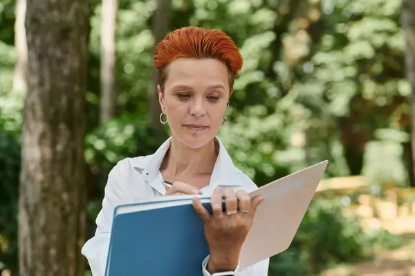 A woman with short, red hair takes notes in a notebook while standing outdoors in a green campus. — Stock Photo