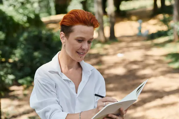 A young woman with bright red hair and a piercing writes in a notebook outdoors. — Stock Photo