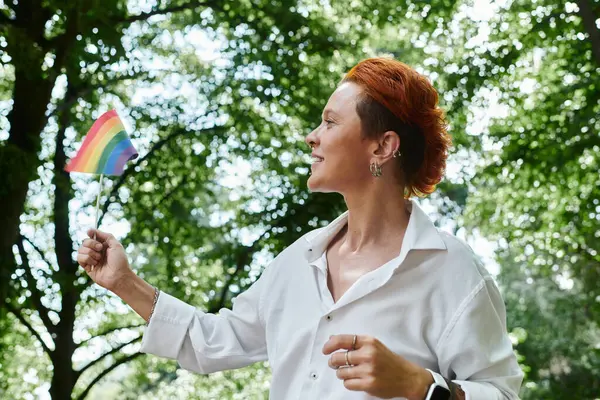 Un giovane insegnante in camicia bianca con i capelli rossi tiene una bandiera di orgoglio arcobaleno tra gli alberi verdi del campus — Foto stock