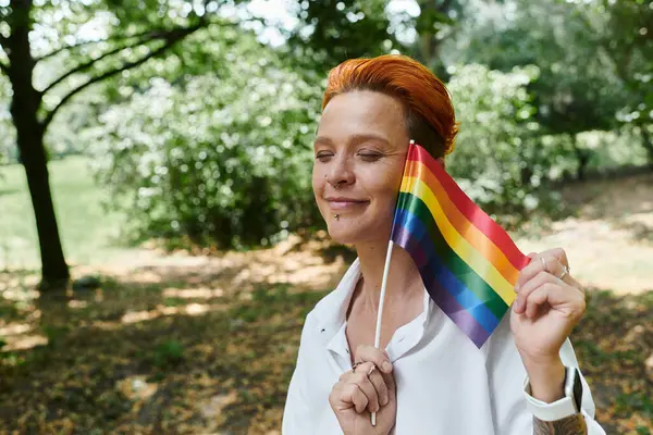 Une enseignante aux cheveux roux courts sourit alors qu'elle tient un drapeau arc-en-ciel dans un parc. — Photo de stock