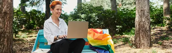 A woman with red hair sits on a park bench working on a laptop, a rainbow flag draped beside her. — Stock Photo