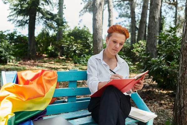 A woman in a white shirt sits on a bench in a campus grove, writing in a red notebook. — Stock Photo