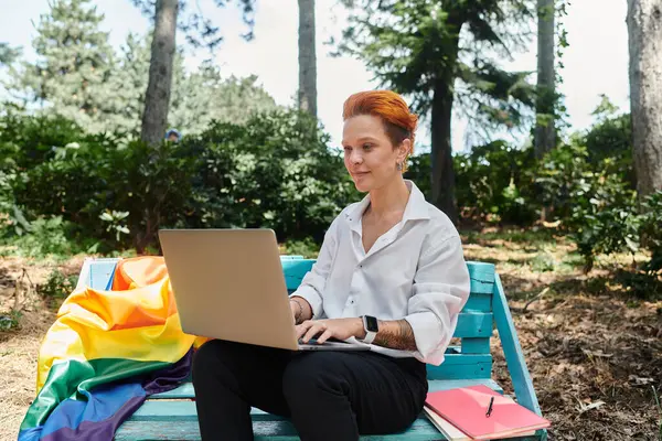 Redhead seated on campus bench uses laptop, rainbow flag in background — Stock Photo