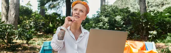 Woman with vivid orange hair sits on campus bench, deep in thought, gazing up from laptop — Stock Photo