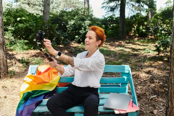 A woman with red hair sits on a bench in a park and takes a selfie with a smartphone. — Stock Photo