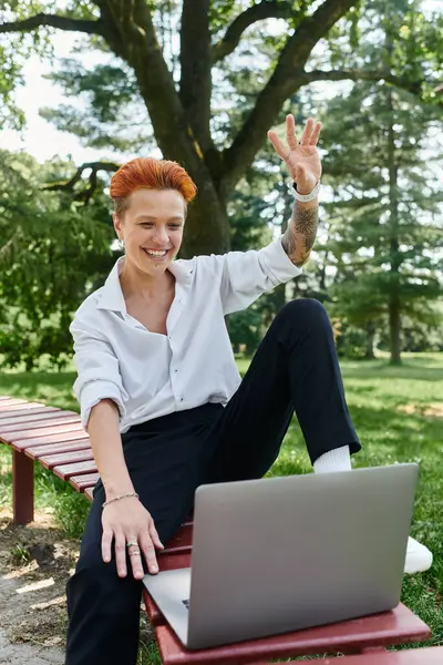 A teacher with orange hair sits on a bench outside, waving and smiling at a laptop. — Stock Photo