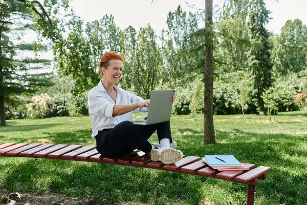 Un insegnante con i capelli rossi sorride mentre usa un computer portatile su una panchina di legno rossa. — Foto stock