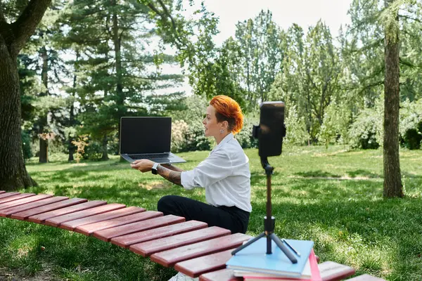 A teacher records a video outdoors, sitting on a bench in a park, showcasing her laptop. — Stock Photo