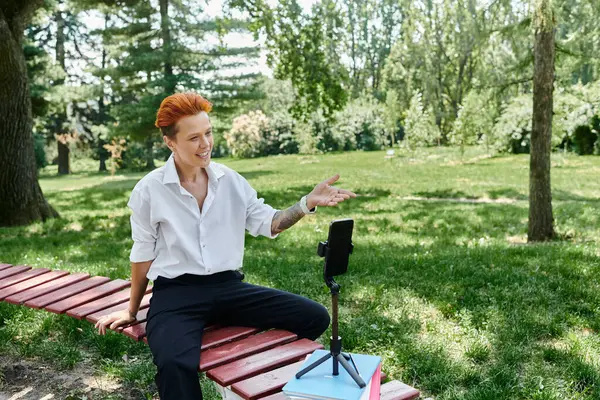 A young teacher sits on a bench in a park, recording a video on her phone. — Stock Photo
