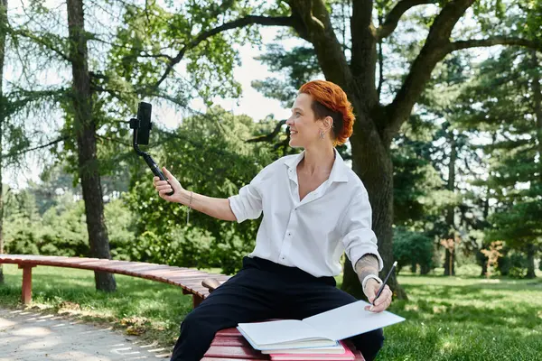 A woman sits on a bench on campus, recording a vlog while holding a phone on a selfie stick. — Stock Photo
