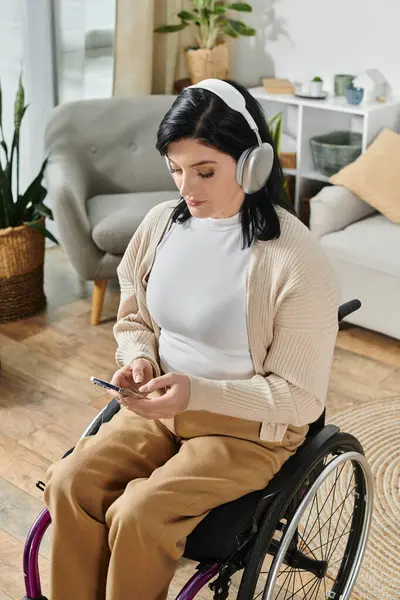 A woman in a wheelchair sits in a room, wearing headphones and looking at a phone. — Stock Photo