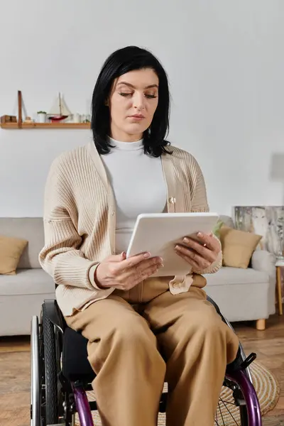 A woman in a wheelchair is seated indoors, looking down at a tablet. — Stock Photo