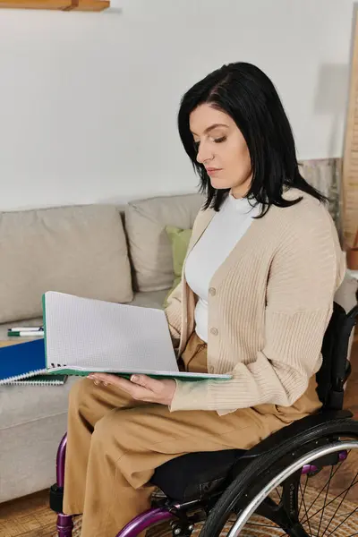 A woman in a wheelchair sits and reads a book in her living room. — Stock Photo