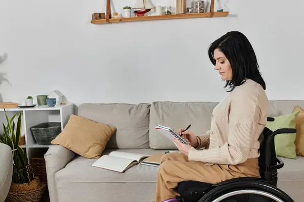 A woman sits in a wheelchair in a living room, writing in a notebook. — Stock Photo