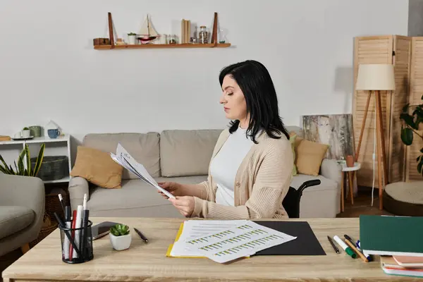 A woman in a wheelchair sits at a desk, reviewing documents in a well-lit living room. — Stock Photo
