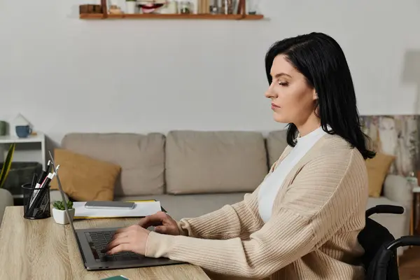 Eine Frau im Rollstuhl sitzt an einem Schreibtisch und tippt auf einem Laptop. — Stockfoto