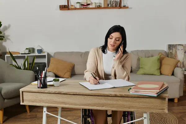 A woman in a wheelchair sits at a desk, taking notes while on a phone call. — Stock Photo