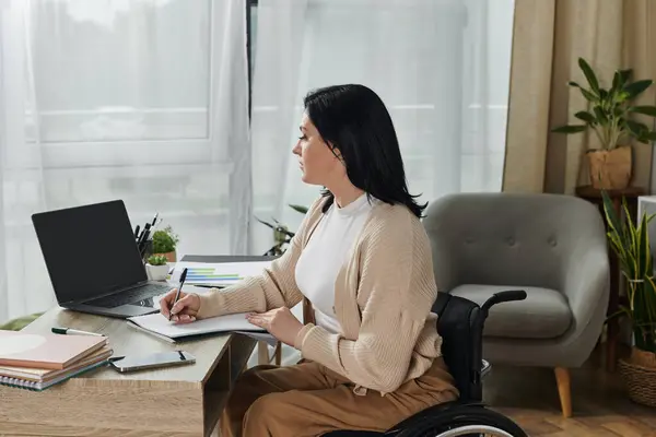 Eine Frau im Rollstuhl sitzt an einem Schreibtisch, arbeitet an einem Laptop und schreibt in ein Notizbuch. — Stockfoto
