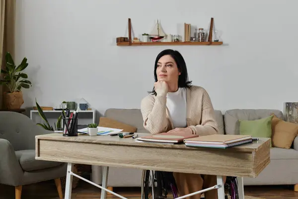 Una donna su una sedia a rotelle si siede a una scrivania nel suo ufficio di casa, guardando pensieroso fuori dal lato. — Foto stock