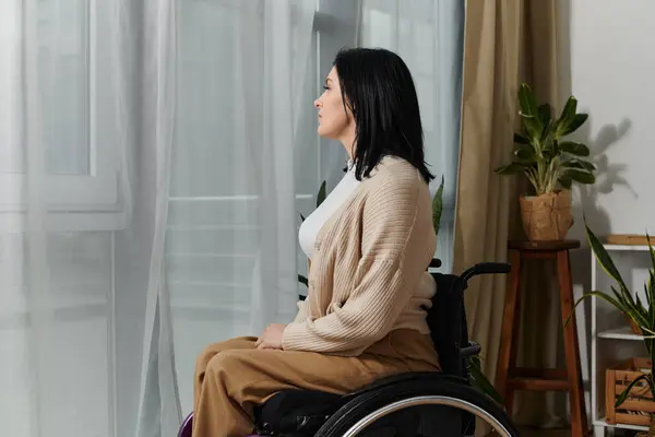 A woman in a wheelchair sits by a window, gazing thoughtfully out. — Stock Photo