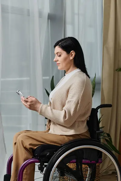 A woman in a wheelchair sits by a window, using her phone. — Stock Photo