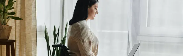 A woman in a wheelchair sits by a window, using a laptop computer. — Stock Photo
