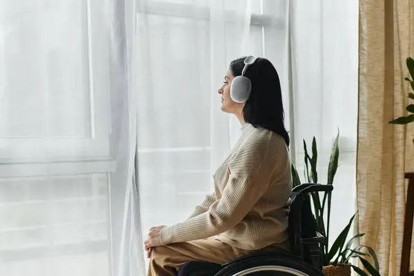 A woman sits in a wheelchair by a window, wearing headphones. — Stock Photo