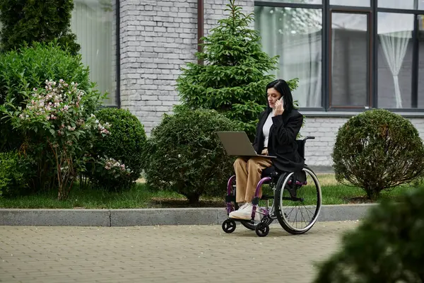 A woman sits in a wheelchair in front of a building, working on a laptop and talking on her phone. — Stock Photo