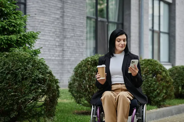 A woman in a wheelchair enjoys a coffee while checking her phone outside an office building. — Stock Photo