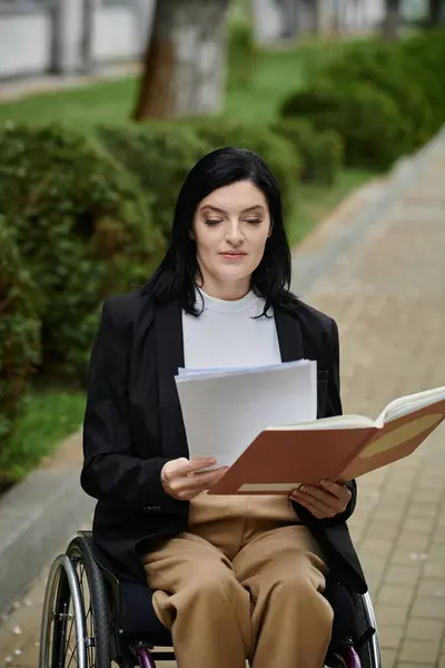 A woman in a wheelchair sits on a paved path in a park and reads documents. — Stock Photo
