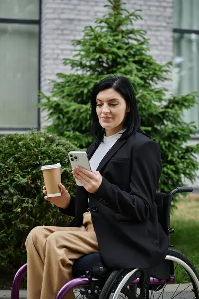 Una mujer en silla de ruedas comprueba su teléfono mientras sostiene una taza de café. - foto de stock