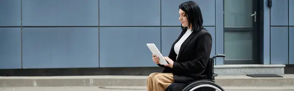 A woman in a wheelchair uses a tablet computer while seated outside a modern building. — Stock Photo