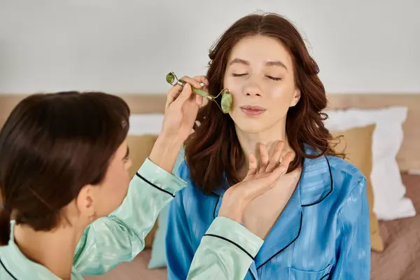 A young woman gently massages her partner face with a jade roller in a cozy bedroom. — Stock Photo