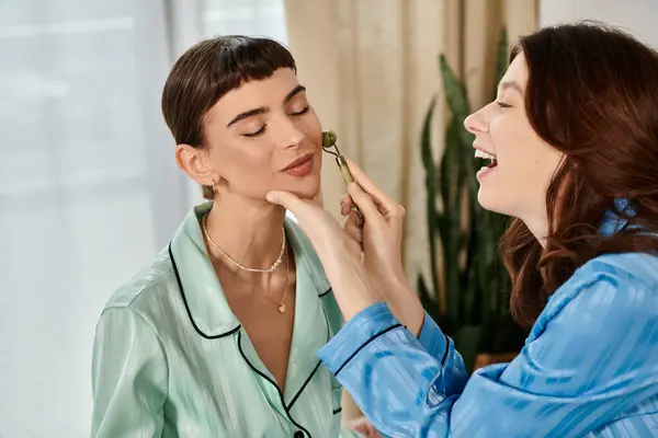 Two women in pajamas, one giving the other a facial massage with a jade roller. — Stock Photo