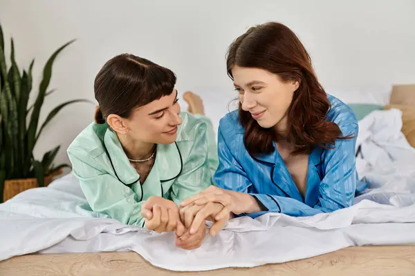 A young lesbian couple, in pajamas, relax and hold hands on a bed, sharing a tender moment. — Stock Photo