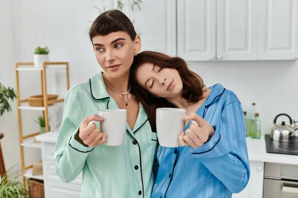 Una joven pareja lesbiana comparte un tranquilo momento matutino, vistiendo pijamas y disfrutando del café en una acogedora cocina. - foto de stock