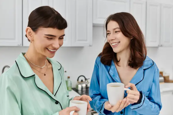 Duas mulheres de pijama, sorrindo e gostando de café juntos na cozinha. — Fotografia de Stock