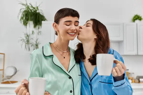 A young couple in pajamas share a tender moment in their kitchen, one woman kissing the other on the cheek while they both hold mugs. — Stock Photo