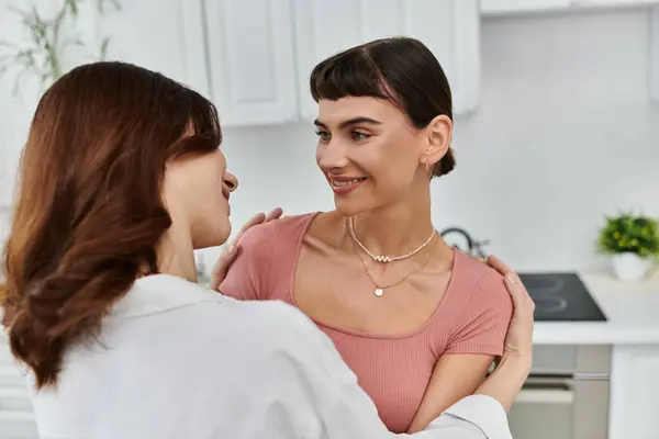 Una giovane coppia lesbica condivide un caldo abbraccio nella loro cucina, il loro amore e la connessione evidente nel loro sguardo. — Foto stock