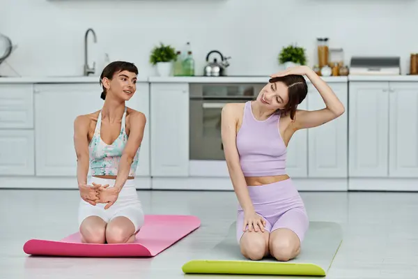 Two young women in athletic wear practice yoga together on mats in a modern kitchen. — Stock Photo