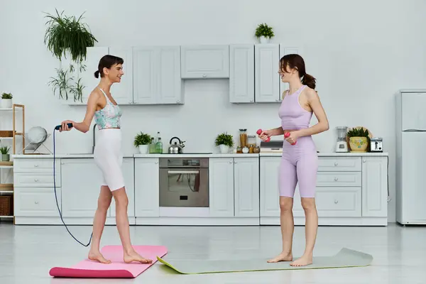 Two women in sportswear work out together in their modern kitchen, one using a jump rope and the other lifting weights. — Stock Photo