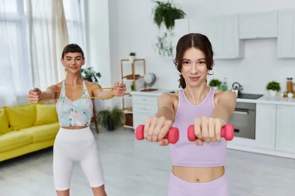 Two young women in sportswear exercise in a modern apartment, one with weights and one with a resistance band. — Stock Photo