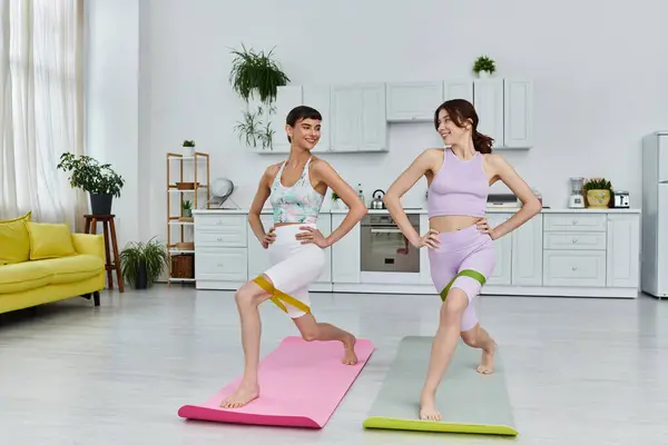 Two women in sportswear work out together in their apartment with resistance bands, smiling and stretching their legs — Stock Photo